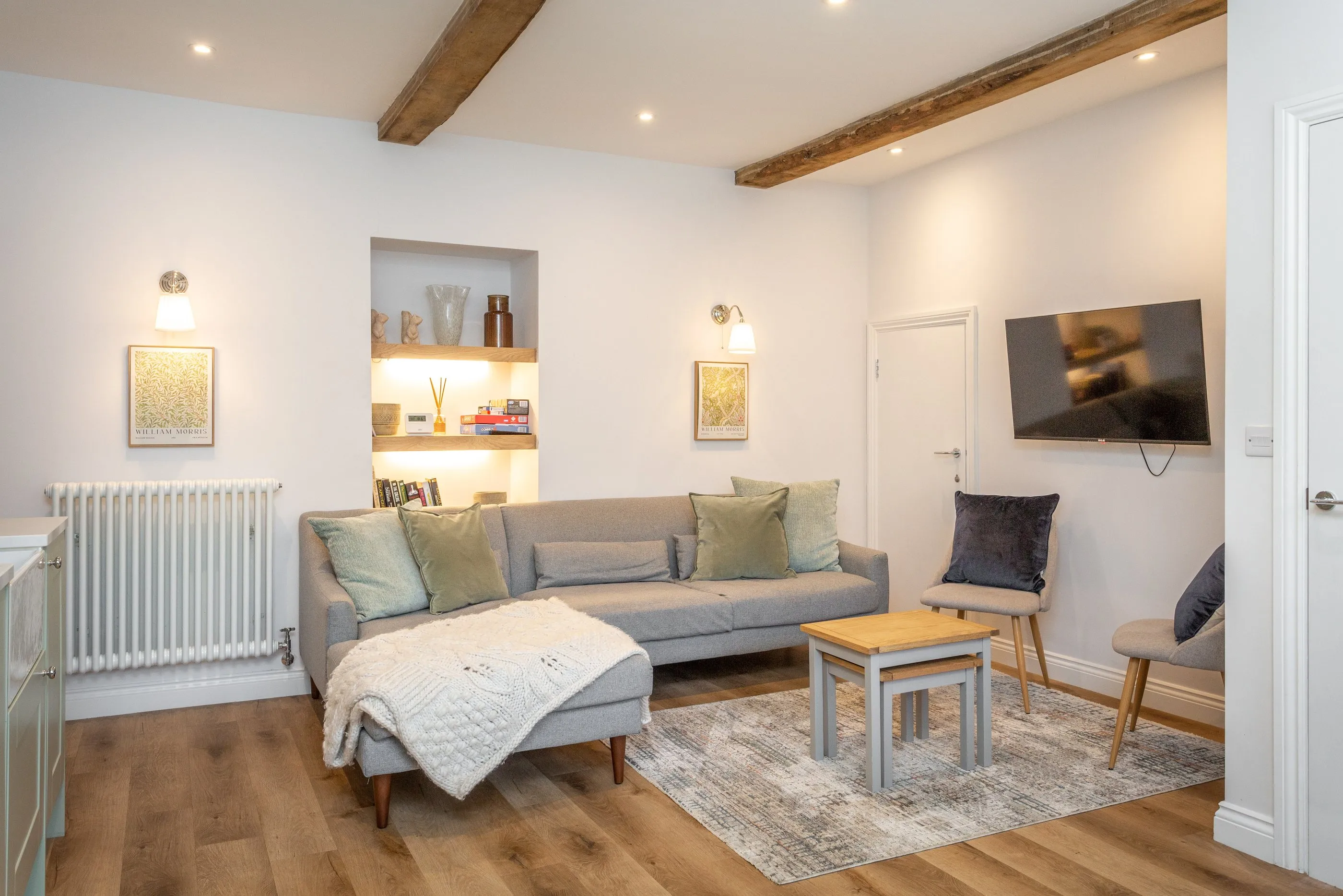Sitting room with oak beams, a grey corner sofa and lit alcove shelves