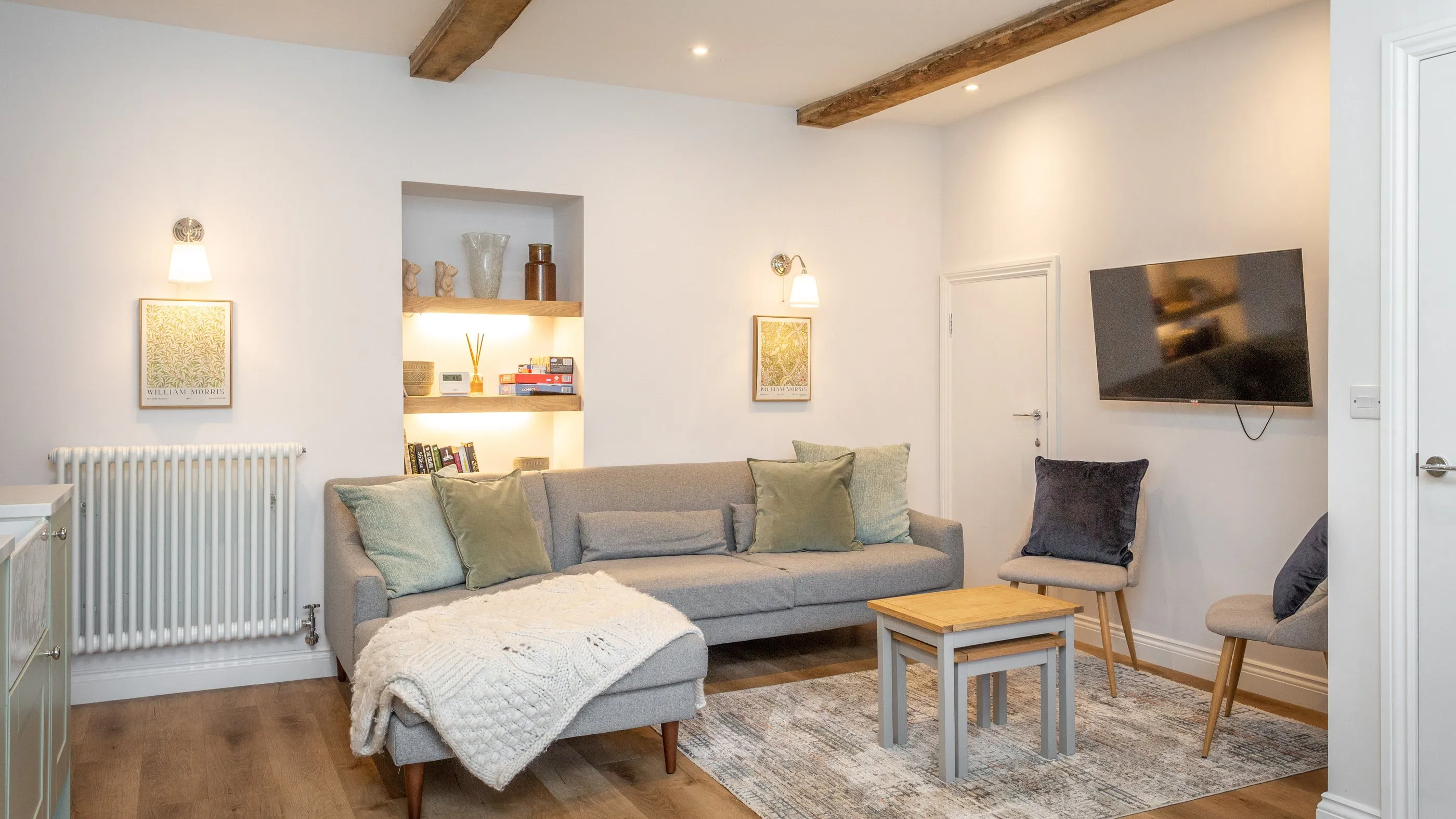 Sitting room with oak beams, a grey corner sofa and lit alcove shelves