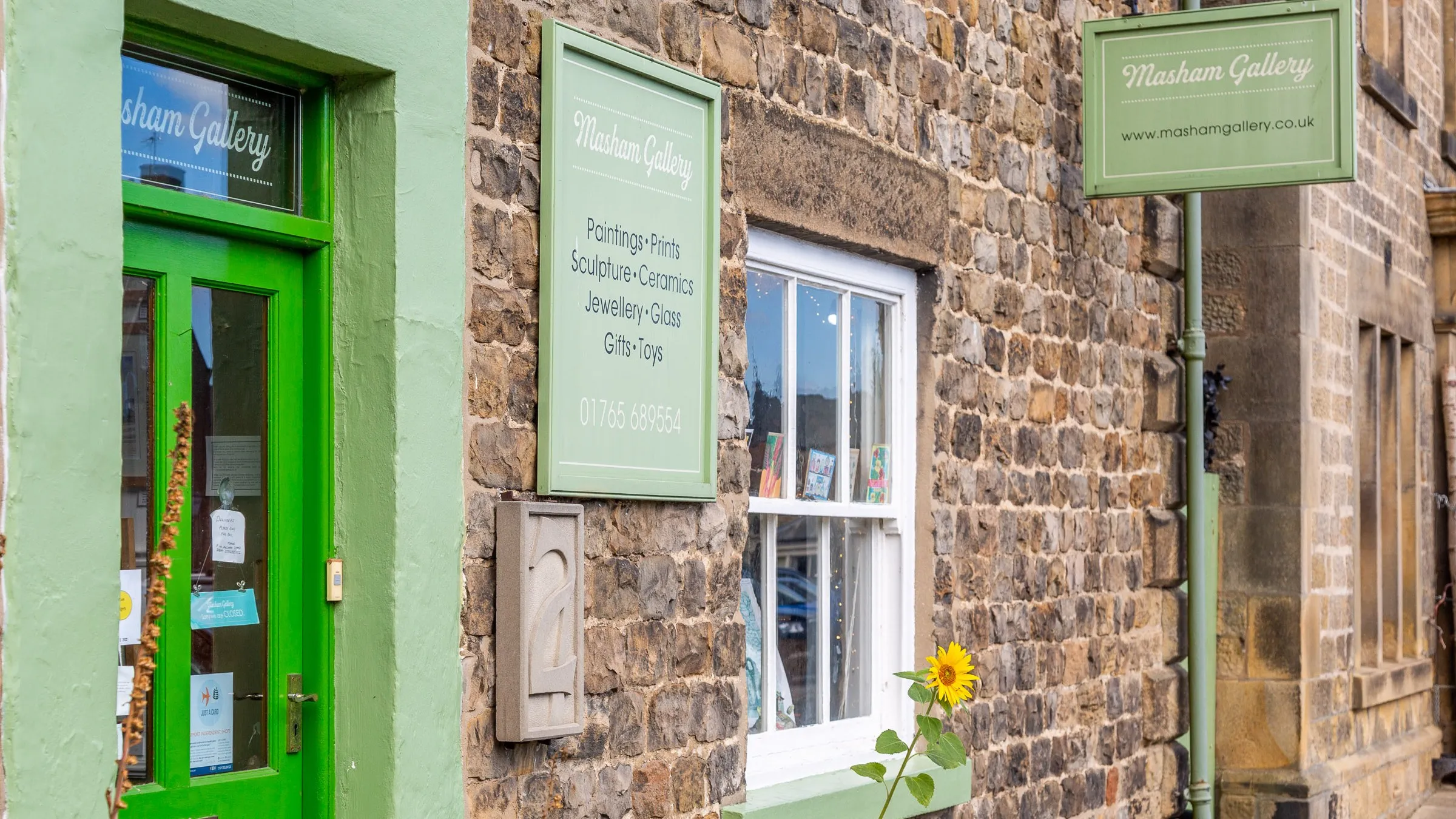 Masham Gallery's green door and stone shopfront, with a sunflower by the window